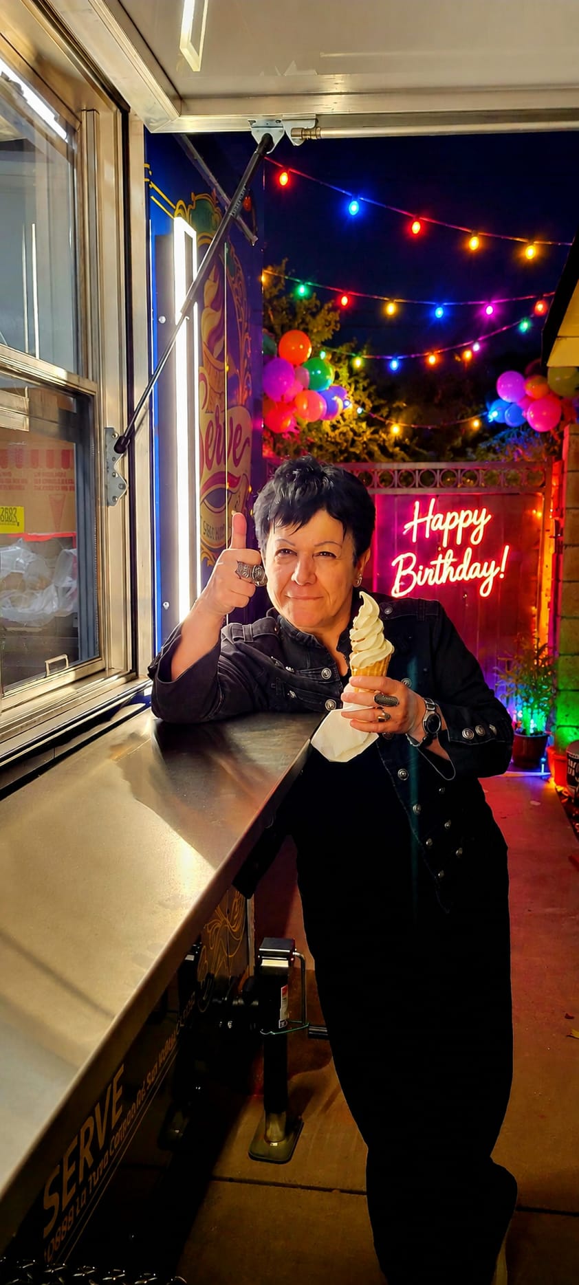 A happy guest gives a thumbs up next to the Serve trailer at a birthday party with string lights and a Happy Birthday neon sign.